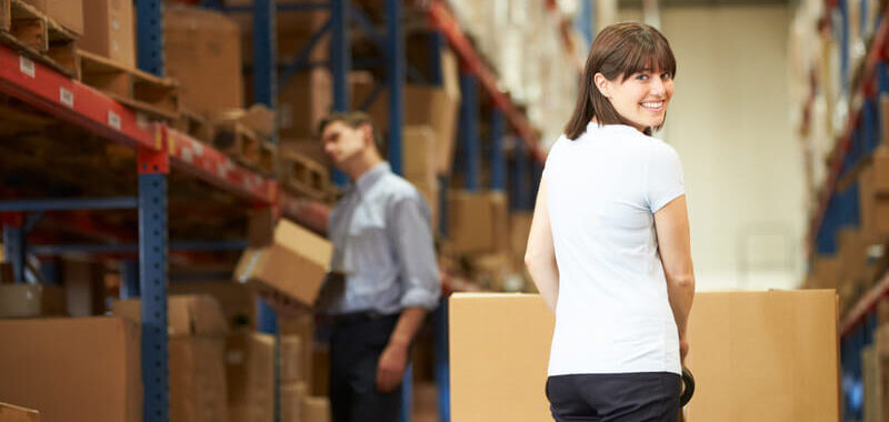 A smiling woman navigating with boxes in a warehouse looks back over her shoulder, with a man looking at warehouse shelves in the background