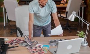 A man stands above desk with printed photos on paper and a laptop.