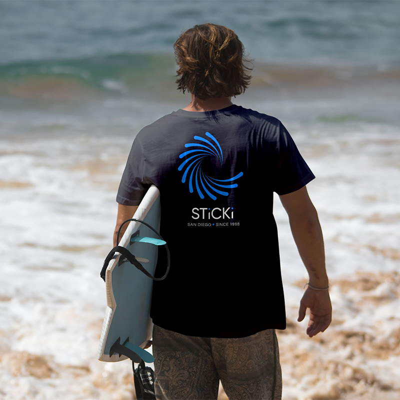 Man with surfboard wearing branded sticki shirt on the beach.